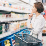 Eine junge Frau steht vor einem Supermarktregal und überlegt, was sie kaufen soll.